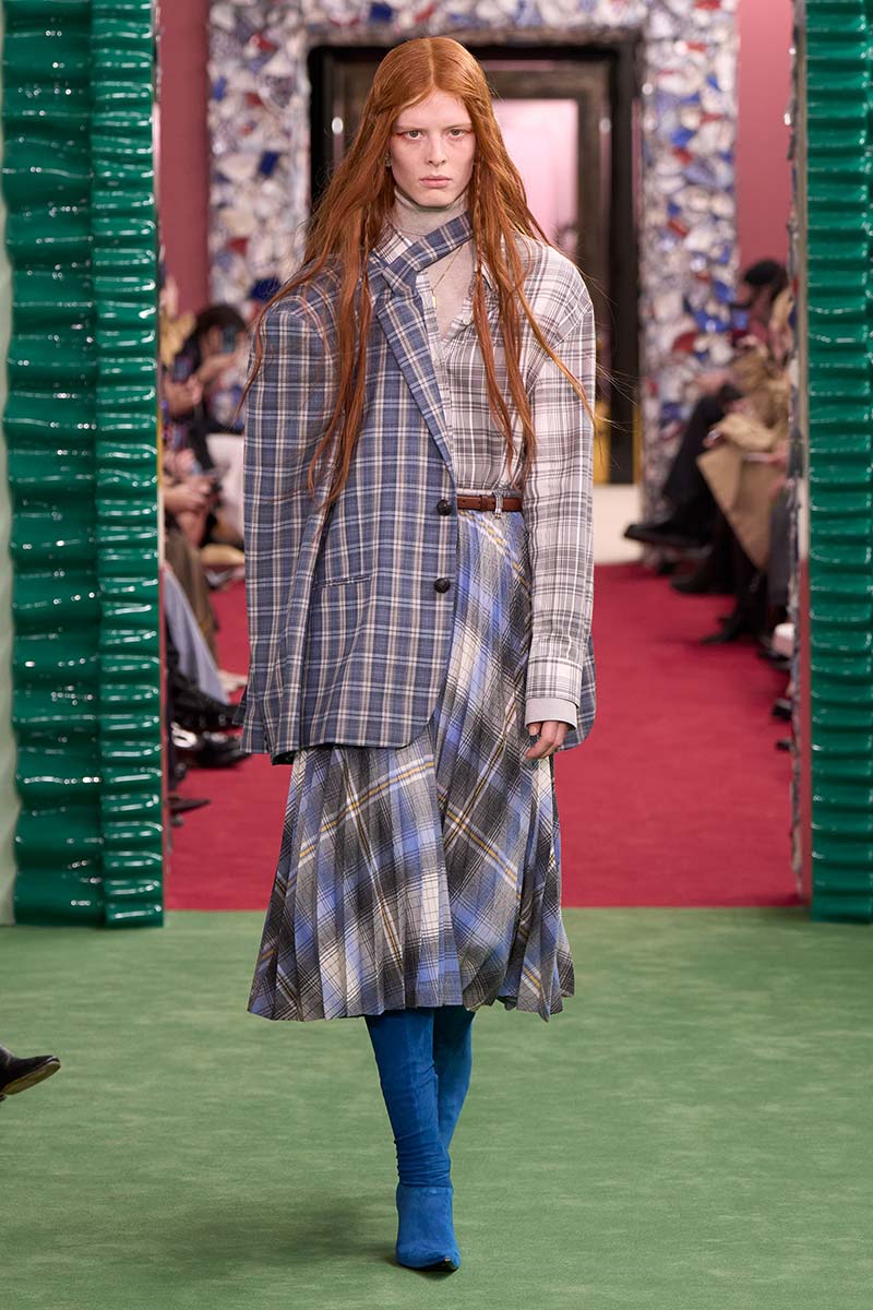 Model op de catwalk in een blazer met Prince of Wales-ruit, een fijn geruite blouse en een geplooide tartan rok in blauw- en grijstinten. Photo courtesy of Acne Studios