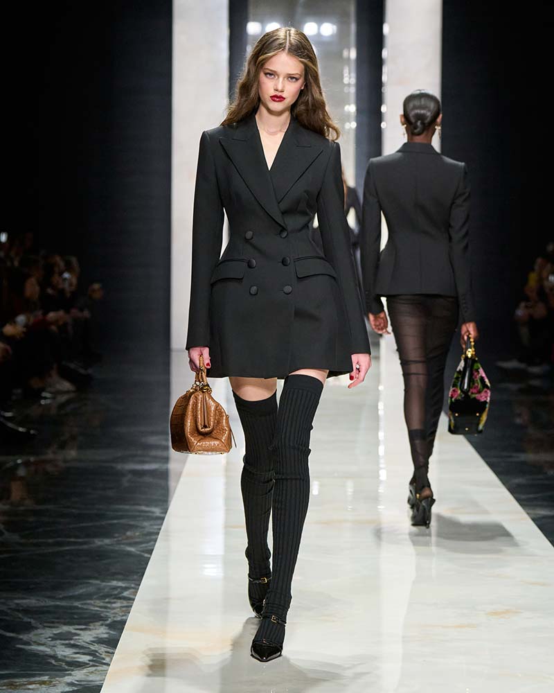 Model in een lange zwarte blazer met hoge zwarte ribkousen en zwarte pumps tijdens de catwalk van Dolce & Gabbana. Photo courtesy of Dolce & Gabbana