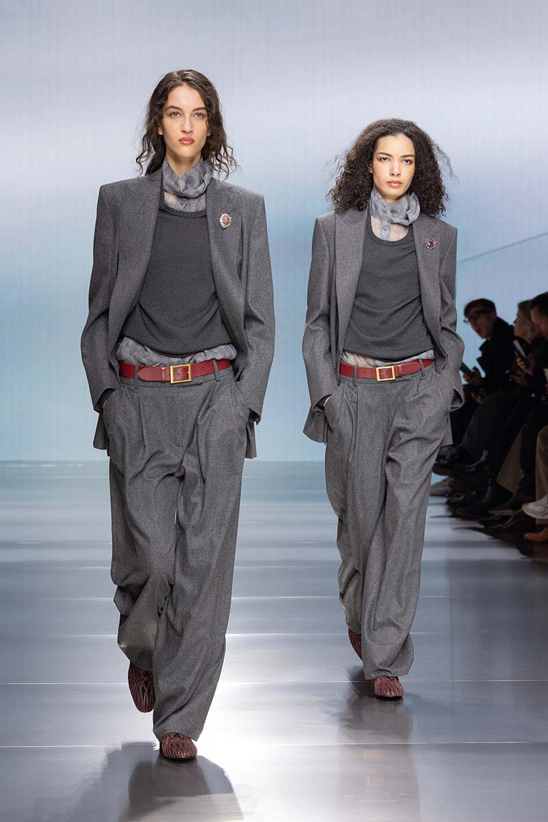 Twee modellen in grijze broekpakken met lichtgrijze blouses en fijngebreide truien op de catwalk van Giorgio Armani. De rode riem geeft de pakken een relaxte twist. Photo courtesy of Giorgio Armani