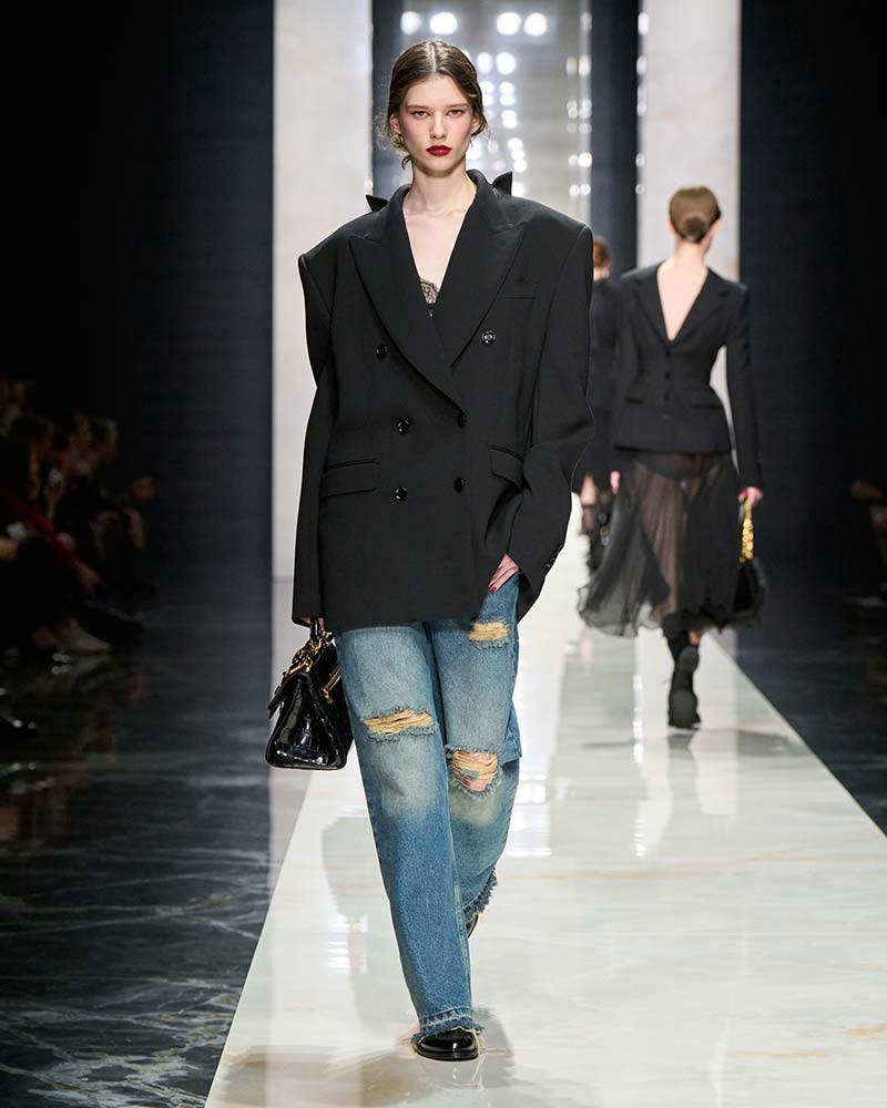 Model draagt een ripped jeans met grote kniegaten, oversized zwarte double-breasted blazer en zwarte platte schoenen met puntige neus. Photo courtesy of Dolce & Gabbana