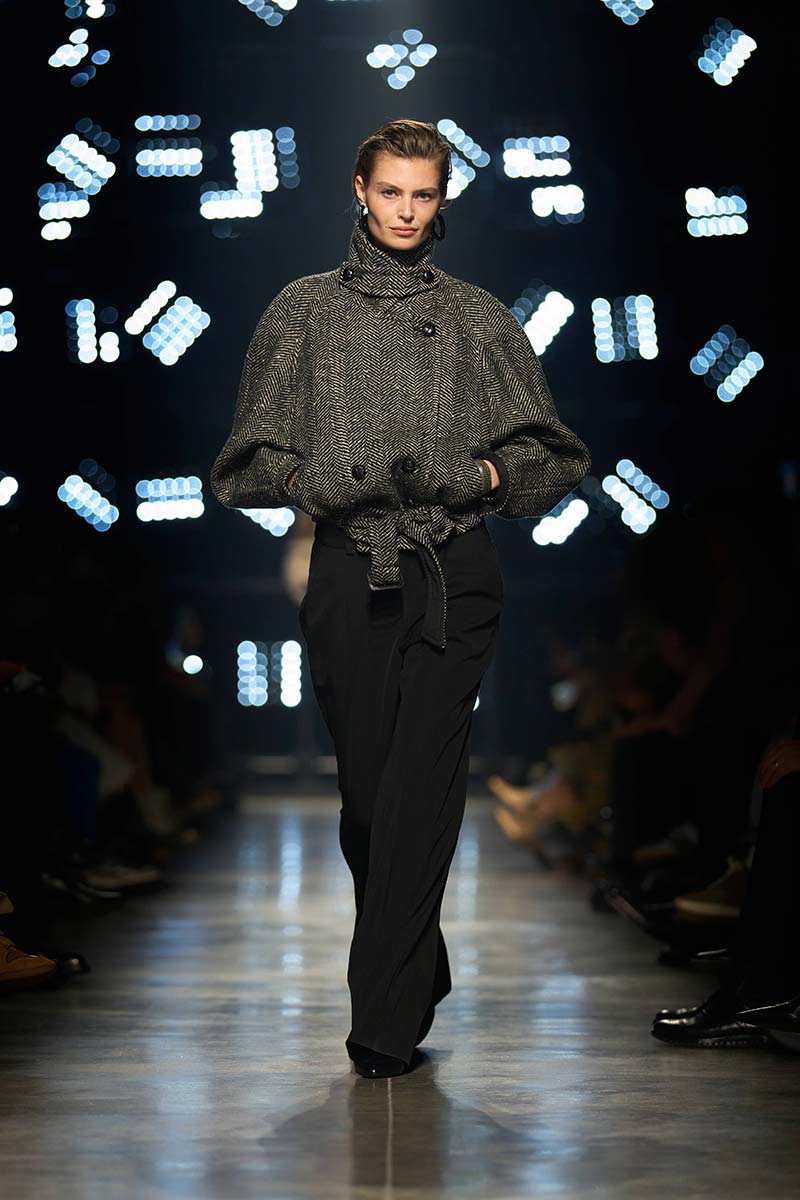 Korte visgraatjas met hoge funnel neck, brede schouders en ceintuur, gecombineerd met een zwarte broek. Photo courtesy of Isabel Marant